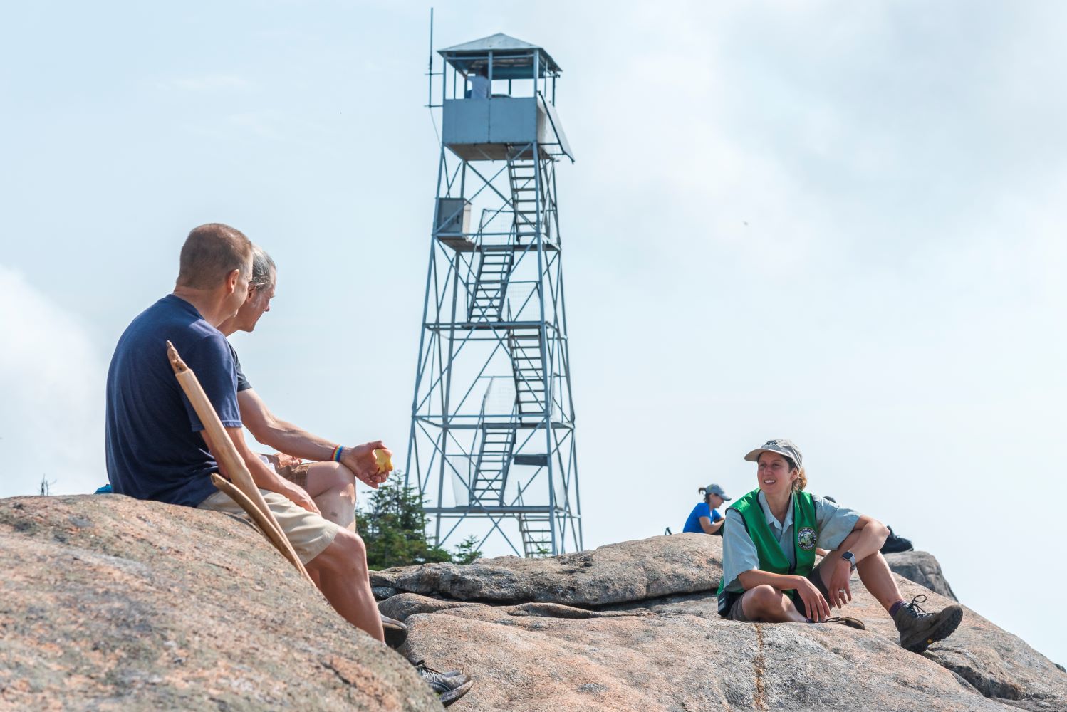 a summit steward speaks to two hikers on a mountain summit with a fire tower standing in the background